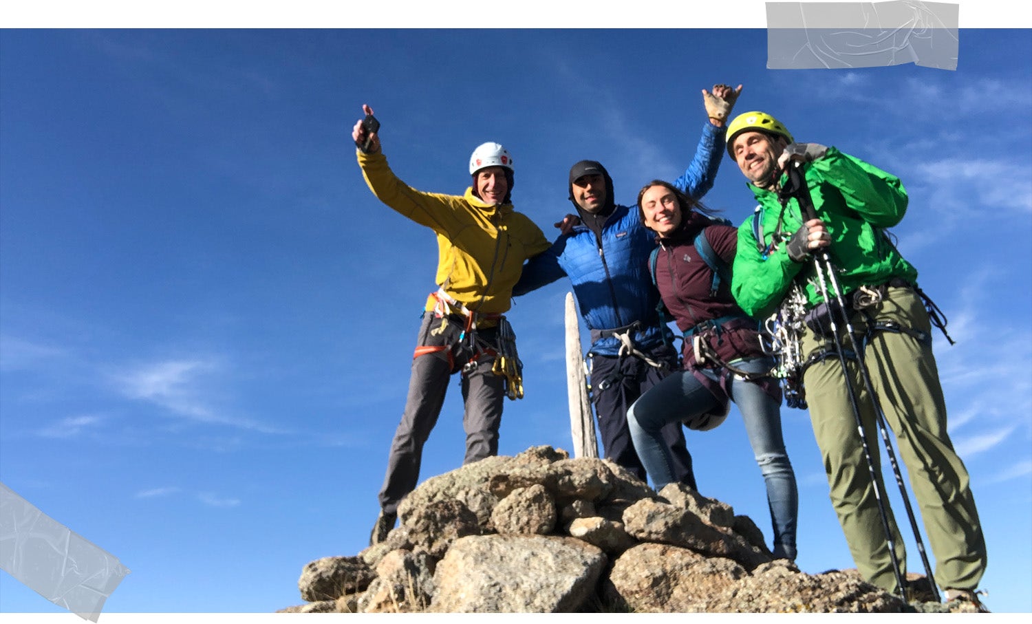 The team smile and wave their arms as they stand on the rocky summit with a blue sky behind them. 