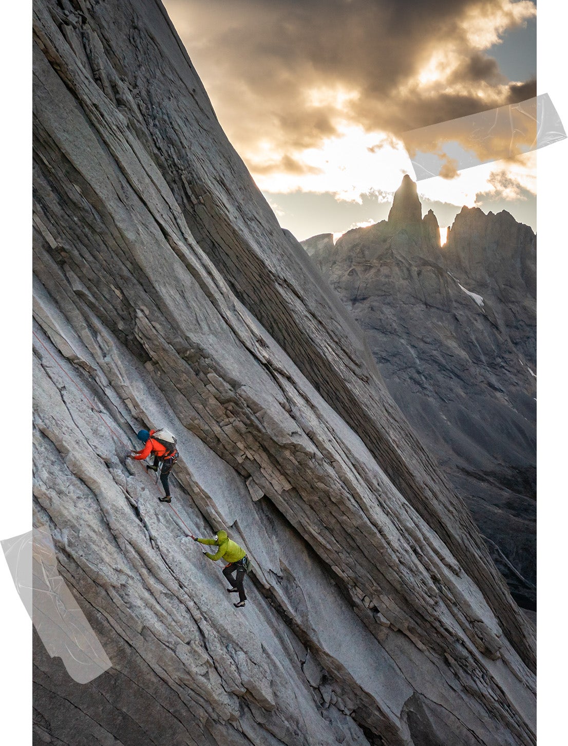 Felipe climbs above Erik on an enormous slab of layered rock at dusk, with the sun peering from behind the jagged mountainous horizon. 