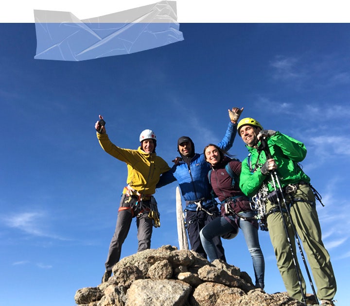 The team smile and wave their arms as they stand on the rocky summit with a blue sky behind them. 