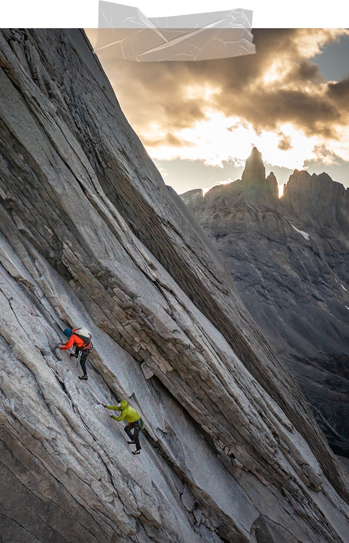 Felipe climbs above Erik on an enormous slab of layered rock at dusk, with the sun peering from behind the jagged mountainous horizon. 